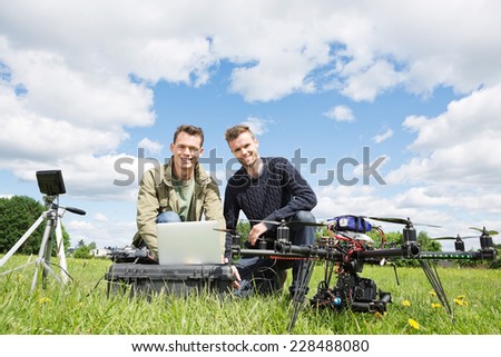 Portrait of men using laptop next to UAV octocopter and tripod against cloudy sky