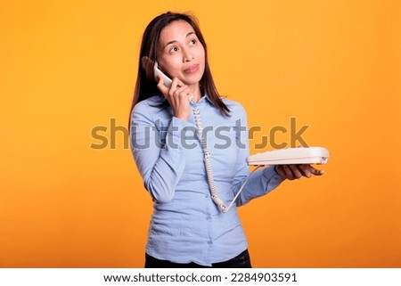 Filipino woman talking on landline phone call, using telephone with cord standing over yellow background. Cheerful model using office phone with cord having remote discussion in studio Royalty-Free Stock Photo #2284903591