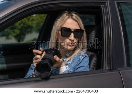 Woman in sunglasses with a camera sits in a car and takes pictures with a professional camera, a private detective or a paparazzi spy. Journalist is looking for sensations and follows celebrities.