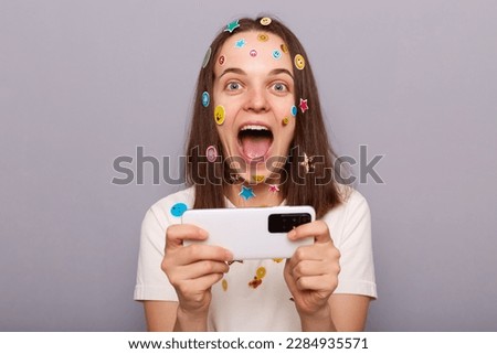 Photo of overjoyed excited woman covered with funny stickers posing isolated over gray background, looking at camera, playing video game, winning online tournament.