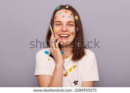 Photo of positive joyful beautiful woman covered with funny stickers holding smartphone and talking with friends, laughing joyfully, posing isolated over gray background
