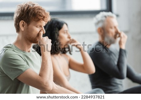 Redhead man practicing nostril breathing near blurred group in yoga studio Royalty-Free Stock Photo #2284937821