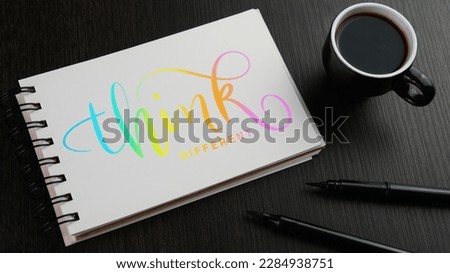 THINK DIFFERENT colorful hand lettering in notebook with cup of coffee and pens on black wooden desk