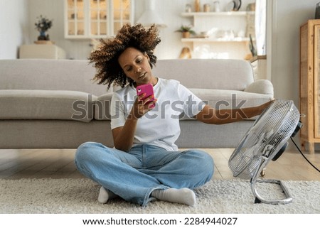 African American girl sitting on floor in living room near electric fan and using smartphone, unhappy black woman cooling down, trying to stay cool without air conditioning, suffering from heat Royalty-Free Stock Photo #2284944027