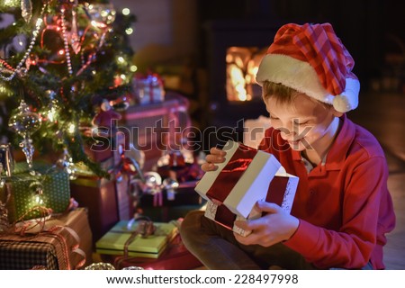 Lovely little boy with a santa claus hat opens a gift in front of the Christmas tree lit up, in the warm atmosphere of Christmas,  a wood stove in the background