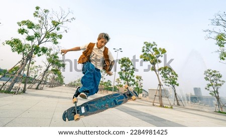 Cool Asian guy skating on longboard skate at park on summer holiday vacation. Stylish young man enjoy and fun urban active lifestyle practicing outdoor sport skateboarding on city street at sunset.