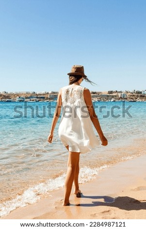 Young woman wearing a white dress and hat standing on a beach and enjoying the sun.