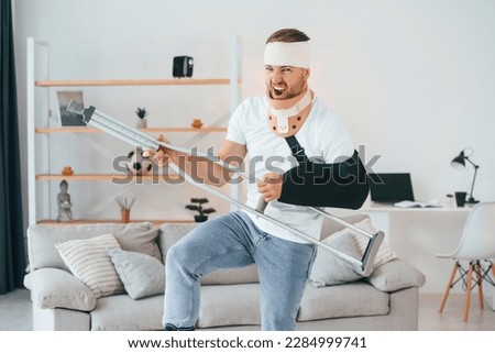Standing, holding crutch and pretending that he's playing guitar. Having fun. Man is at home indoors. Having injuries. Royalty-Free Stock Photo #2284999741