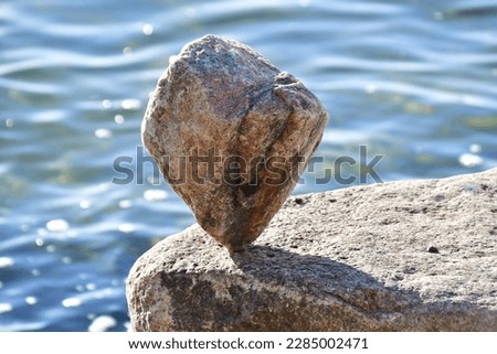 A kite-shaped stone balances on its top on a rock by the water