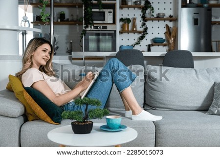 Young freelancer woman wedding planner and counselor sitting on sofa drinking coffee and coming up with new ideas for clients as she reads and write what she has written from the wedding plans options