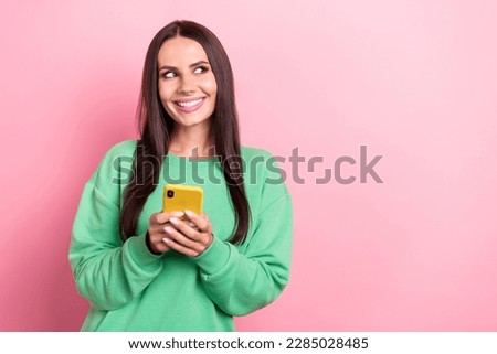 Photo of cheerful satisfied woman dressed green sweatshirt look at empty space holding smartphone isolated on pink color background