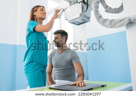 Male patient lying on bed while female nurse adjusting modern X-ray machine for scanning his arm or hand for injuries and fractures