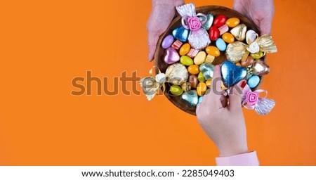 Serving candies, mother holding bowl and serving candies. Top view isolated orange background, copy space. Ramadan or Ramazan feast celebration concept idea. Girl hand taking one wrapped chocolate. Royalty-Free Stock Photo #2285049403