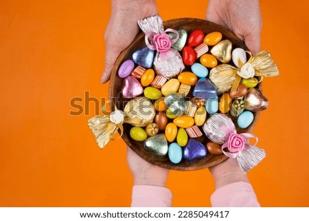 Holding bowl of sweet, woman and child hands holding bowl of sweet. Top view image of wrapped luxury chocolate and almond dragee in wooden tray. Isolated orange studio background with copy space. Royalty-Free Stock Photo #2285049417