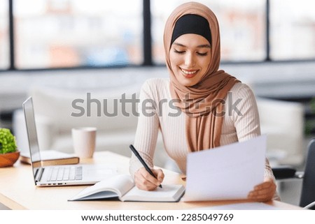 beautiful young smiling muslim woman in traditional religious hijab works remotely on laptop from home
