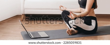 woman doing yoga at home sitting on mat holding fingers and watching tutorial lesson on laptop