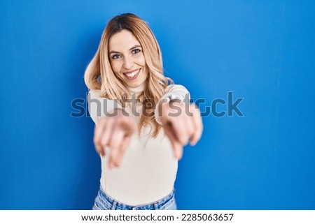 Young caucasian woman standing over blue background pointing to you and the camera with fingers, smiling positive and cheerful 