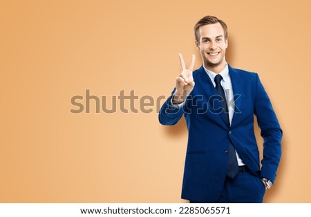 Excited businessman in confident suit showing two fingers or victory sign gesture, over latte beige background. Happy gesturing man. Copy space for ad slogan or text.