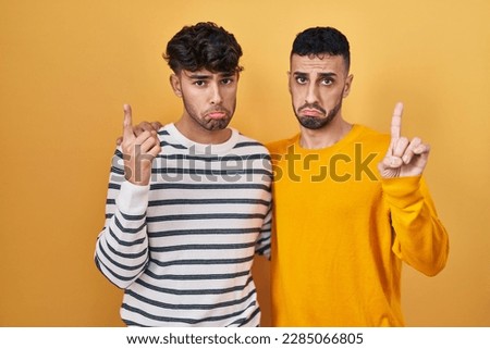 Young hispanic gay couple standing over yellow background pointing up looking sad and upset, indicating direction with fingers, unhappy and depressed. 