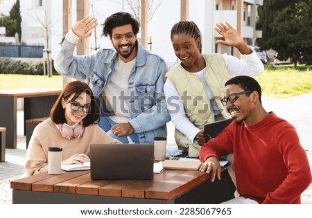Smiling young multiethnic people students studying with laptop together, wave hand, say hi, greeting teacher outdoor. Modern education with device, group lesson, project work in college Royalty-Free Stock Photo #2285067965
