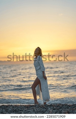 Attractive young elegant lady in dress enjoying sunset time at the beach. Background orange sundown on the sea
