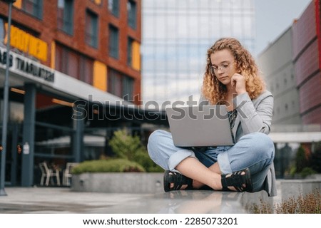Photo of beautiful curly haired woman sitting outdoors in front of university campus holding notebook typing essay text wearing eyeglasses. Online communication