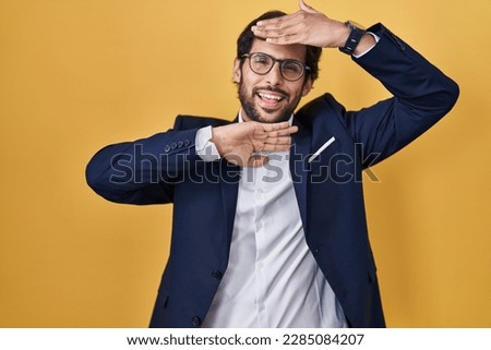 Handsome latin man standing over yellow background smiling cheerful playing peek a boo with hands showing face. surprised and exited 