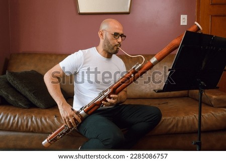 young latino man, bald with glasses and a white shirt, is sitting on a leather sofa playing the bassoon, reading and studying sheet music at home. Royalty-Free Stock Photo #2285086757