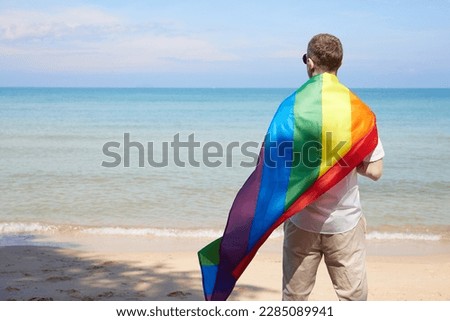 back young man looking to the sea and holding a rainbow flag(LGBT) on a tropical beach