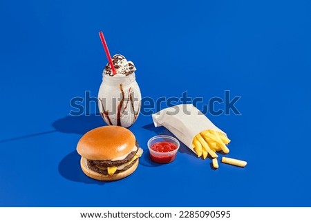Tempting combo meal with burger, fries, sauce, and milkshake on a blue background. Minimalist style showcases fast food in a modern, unhealthy way. Royalty-Free Stock Photo #2285090595