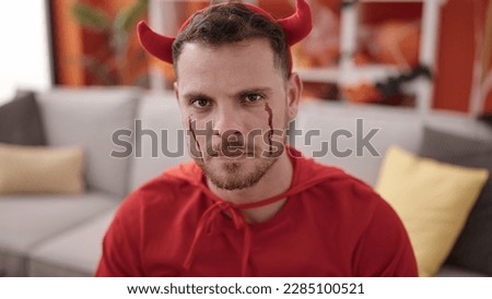 Young caucasian man wearing devil costume sitting on the sofa at home