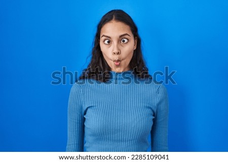 Young brazilian woman standing over blue isolated background making fish face with lips, crazy and comical gesture. funny expression. 