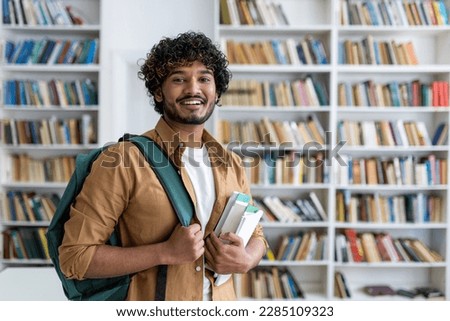 Portrait of happy satisfied african american student with curly hair, guy with books and backpack standing in university academic library smiling and looking at camera.