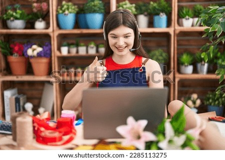 Young caucasian woman working at florist shop doing video call smiling happy and positive, thumb up doing excellent and approval sign 