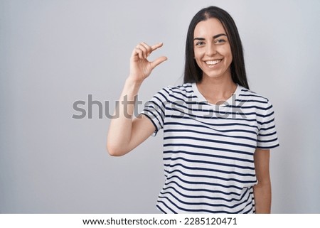 Young brunette woman wearing striped t shirt smiling and confident gesturing with hand doing small size sign with fingers looking and the camera. measure concept.  Royalty-Free Stock Photo #2285120471