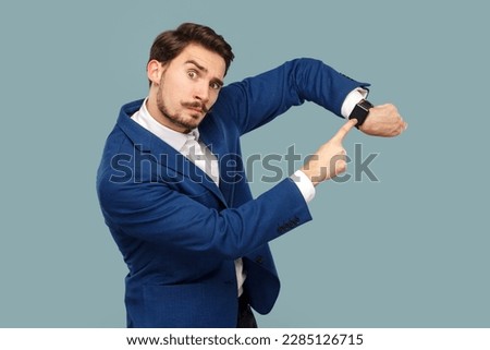 Portrait of serious assertive man with mustache standing pointing at his wristwatch, saying you have no time, wearing white shirt and jacket. Indoor studio shot isolated on light blue background. Royalty-Free Stock Photo #2285126715