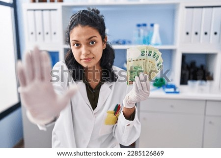 Hispanic woman with dark hair working at scientist laboratory holding money with open hand doing stop sign with serious and confident expression, defense gesture 