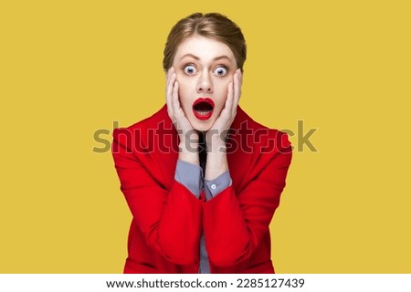 Portrait of amazed surprised woman with red lips with hands on cheeks, looking at camera with big eyes, sees something scary, wearing red jacket. Indoor studio shot isolated on yellow background.