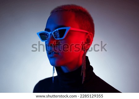 Portrait of a young woman with a short male haircut in sunglasses in neon light in the studio