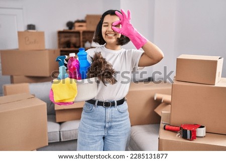 Young hispanic woman cleaning at new home smiling happy doing ok sign with hand on eye looking through fingers 