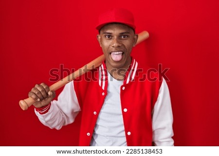 Young hispanic man playing baseball holding bat sticking tongue out happy with funny expression. emotion concept. 