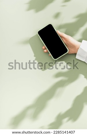 Woman hold phone with black blank screen, minimal flat lay on olive green background. Smartphone mock up. Digital online summer lifestyle composition with leaves shadows and sunlight, pastel colors