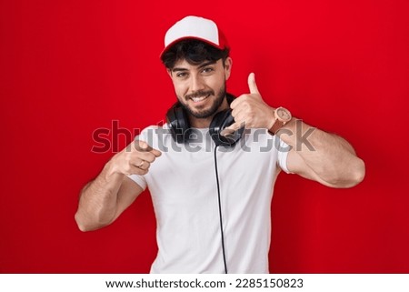 Hispanic man with beard wearing gamer hat and headphones smiling doing talking on the telephone gesture and pointing to you. call me. 