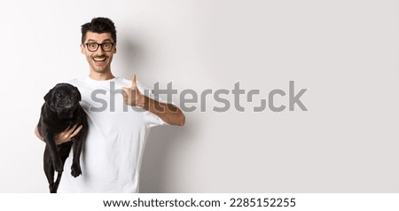 Cheerful handsome man holding dog and showing okay sign, approve or recommend product. Hipster guy carry cute black pug and looking satisfied, white background.