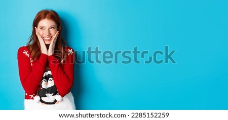 Winter holidays and Christmas Eve concept. Adorable redhead girl blushing and touching cheeks from happiness, smiling and looking at upper left corner promo, blue background.