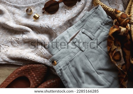 Vintage sweater, neutral trousers, wicker bag, leopard scarf, brown shoes, sunglasses and brass rings on wooden background. Top view.