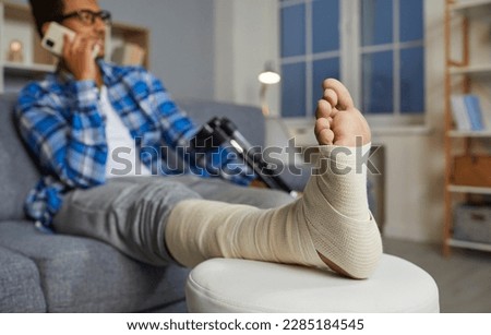 Close up of an injured foot. African American man talking on the mobile phone while relaxing on the sofa at home, with his foot and leg in a bandage resting on a stool. Physical injury concept Royalty-Free Stock Photo #2285184545