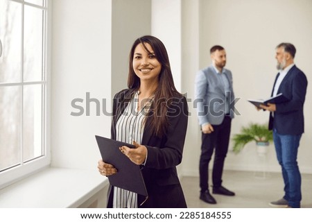 Portrait friendly female business consultant, secretary, estate agent, assistant manager at work. Happy beautiful smiling young woman with clipboard standing in office with other workers in background Royalty-Free Stock Photo #2285184555