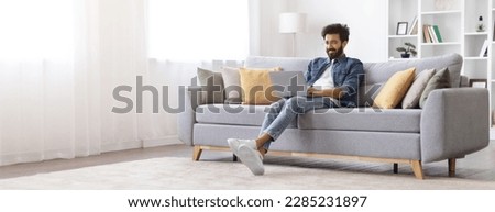 Remote Work. Smiling Indian Freelancer Guy Working With Laptop At Home, Millennial Eastern Man Sitting On Couch In Living Room And Using Computer, Enjoying Distance Job, Panorama With Copy Space Royalty-Free Stock Photo #2285231897