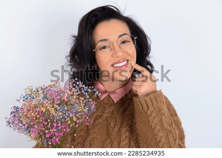 Happy beautiful woman wearing knitted sweater over white background with toothy smile, keeps index fingers near mouth, fingers pointing and forcing cheerful smile